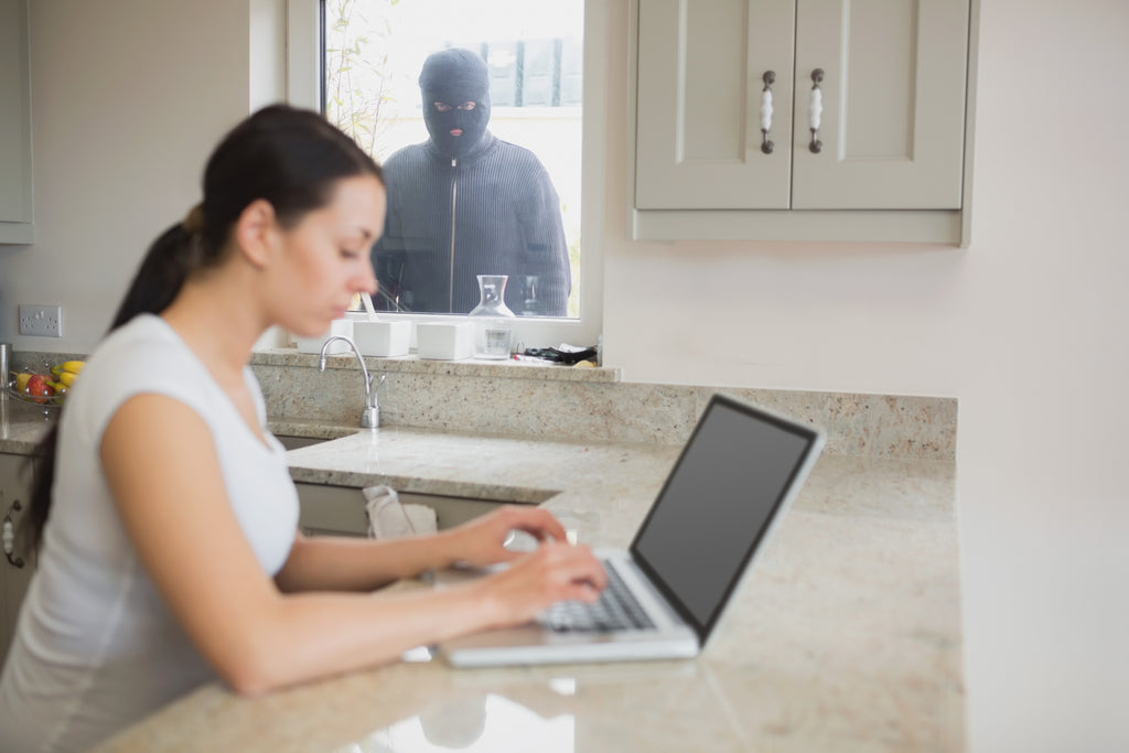 Eine Frau, die in ihrer hellen Küche auf ihren Laptop konzentriert ist, bemerkt die maskierte Gestalt draußen nicht, die durchs Fenster schaut. Das Bild symbolisiert im Zusammenhang mit IDEAL Aktenvernichtern die Notwendigkeit von Datenschutz im Sinne des Schutzes sensibler Daten auf Papier im Homeoffice. 