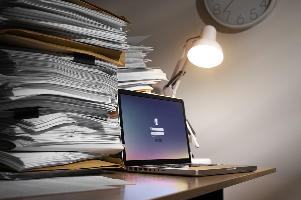 Ein unordentlicher Schreibtisch mit zwei großen Stapeln Papier neben einem offenen Laptop und einer eingeschalteten Schreibtischlampe. Eine analoge Wanduhr ist teilweise oben sichtbar.