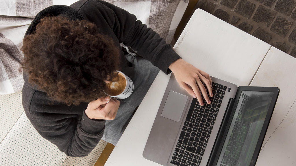 Die Draufsicht zeigt eine Person mit lockigem Haar in ihrem Homeoffice, mit der Hand auf einem Laptop, die gerade Kaffee aus einer Tasse trinkt. Ein weißer Tisch, ein Ziegelboden und Textilien runden die gemütliche Szene ab und betonen sowohl Produktivität als auch Privatsphäre. Es geht um Datenschutz im Zusammenhang mit IDEAL Aktenvernichtern - ohne dass ein Gerät zu sehen ist.