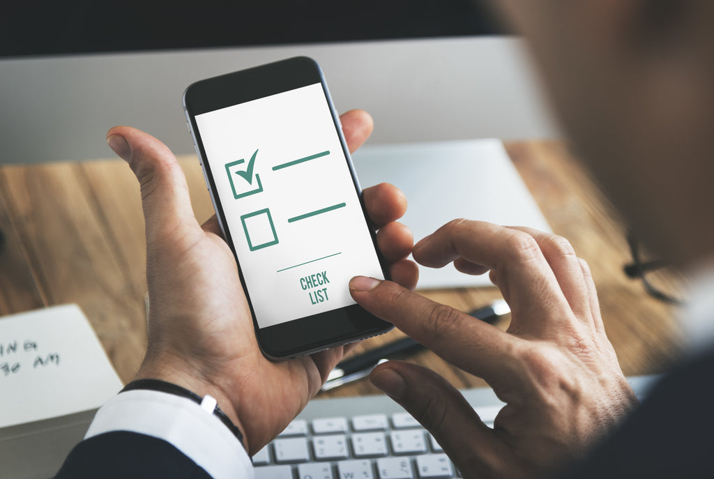 Eine Hand hält ein Smartphone, auf dem eine kurze Checkliste mit einem aktivierten und zwei deaktivierten Kästchen angezeigt wird, auf dem Schreibtisch dahinter sind eine Tastatur und Notizen auf Papier zu sehen.