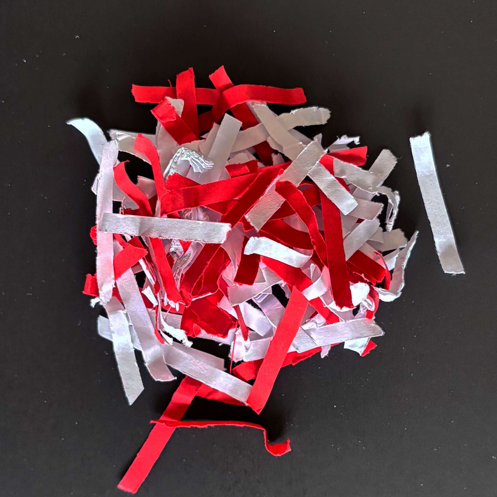 Geshredderte Papierschnipsel in weiß-rot der Sicherheitsstufe P4 - im Zusammenhang mit IDEAL Aktenvernichtern, schlicht vor grauem Hintergrund.