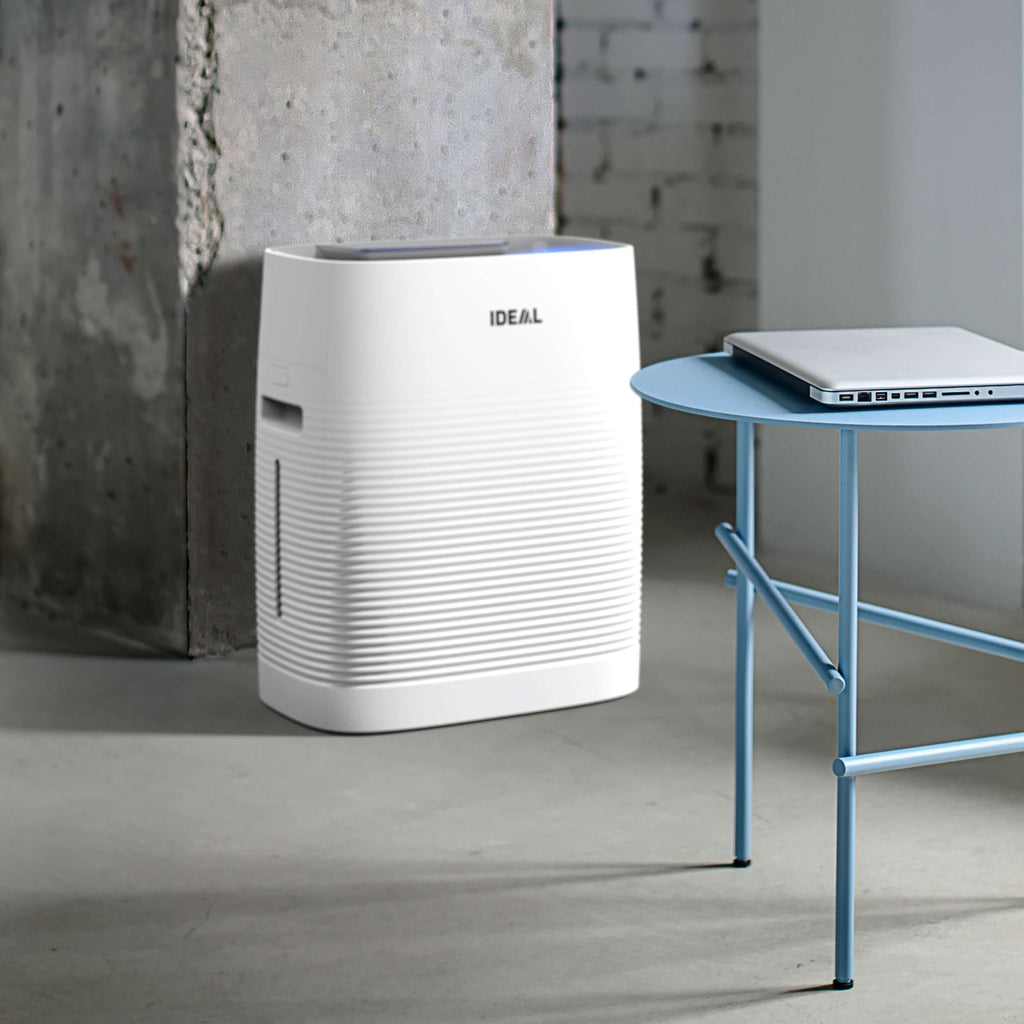 Weißer Luftreiniger der Marke IDEAL steht neben einem blauen Beistelltisch mit geschlossenem Laptop darauf vor einer Betonwand.