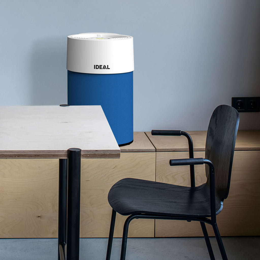 Minimalistisches Büro in Grautönen. Blick auf einen Stuhl und einen teilweise sichtbaren modernen Schreibtisch.
Dahinter an der Wand ein IDEAL AP 30 PRO Luftreiniger auf einer breiten Holzbank.