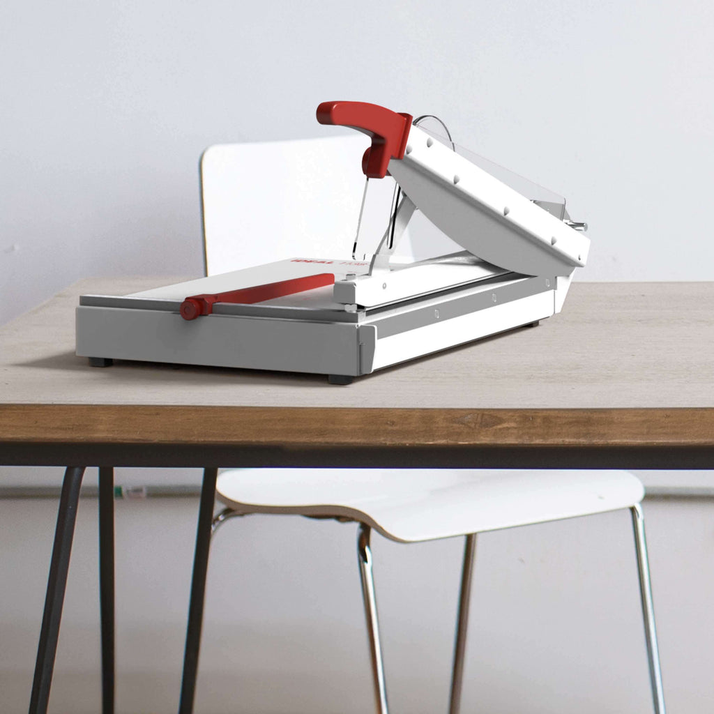 Auf einem modernen, hölzernen Schreibtisch steht ein professioneller Hebelschneider in hellgrau-rot - das Modell IDEAL 1138. Hinter dem Tisch ein schlichter weißer Stuhl.