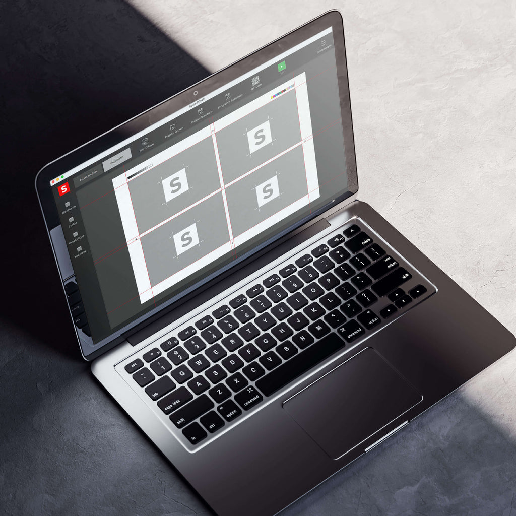 Ein Laptop auf einer grauen Oberfläche zeigt die Oberfläche der Speedcut-Schnittautomatisierungs-Software von IDEAL THE 56-Stapelschneidern mit vier identischen quadratischen Designs, die den Buchstaben S zeigen und an einen Druckbogen erinnern. Sonnenlicht wirft Schatten auf den Laptop und die Oberfläche.