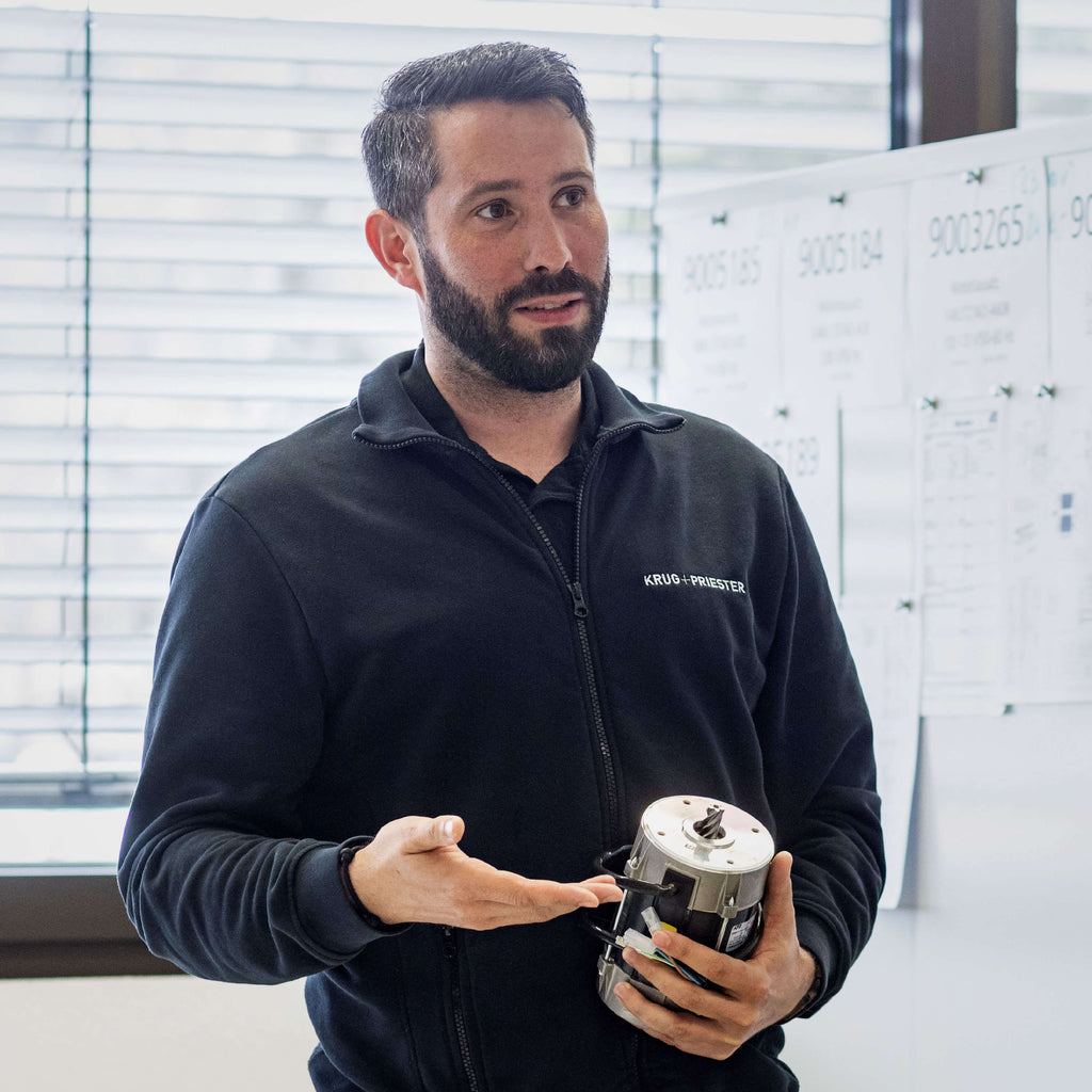 Ein Mann in schwarzem Pullover hält einen Werkstück in der Hand. Im Hintergrund sind Dokumente an einer Wand sichtbar.