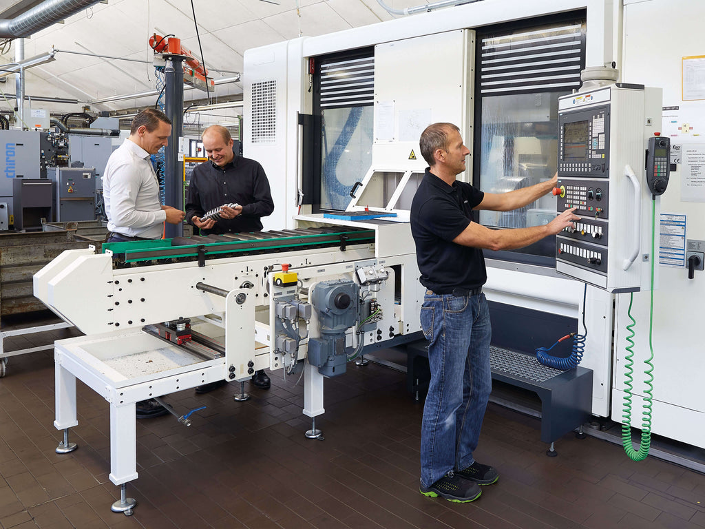 Drei Personen in einer Produktionsumgebung. Eine Person in schwarzem Shirt bedient eine Maschine, während zwei andere Personen mit einem Gerät in der Hand stehen und die Maschine beobachten. Im Hintergrund sind Maschinen und Werkbänke sichtbar.