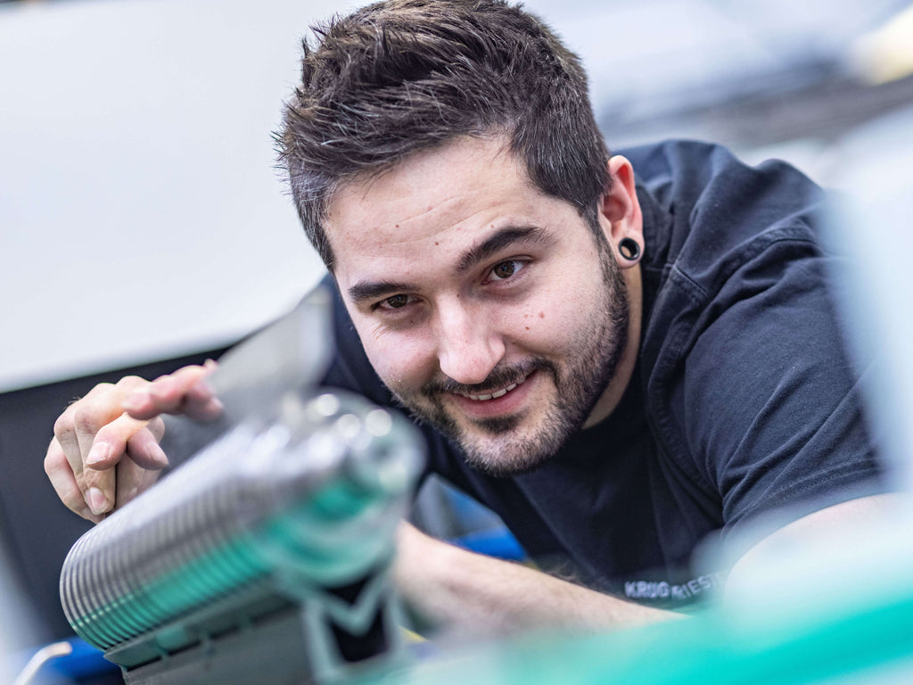 Ein Mann mit kurzen, dunklen Haaren kontrolliert mit einem Messchieber eine Messerwelle. Er trägt ein schwarzes T-Shirt und lächelt.