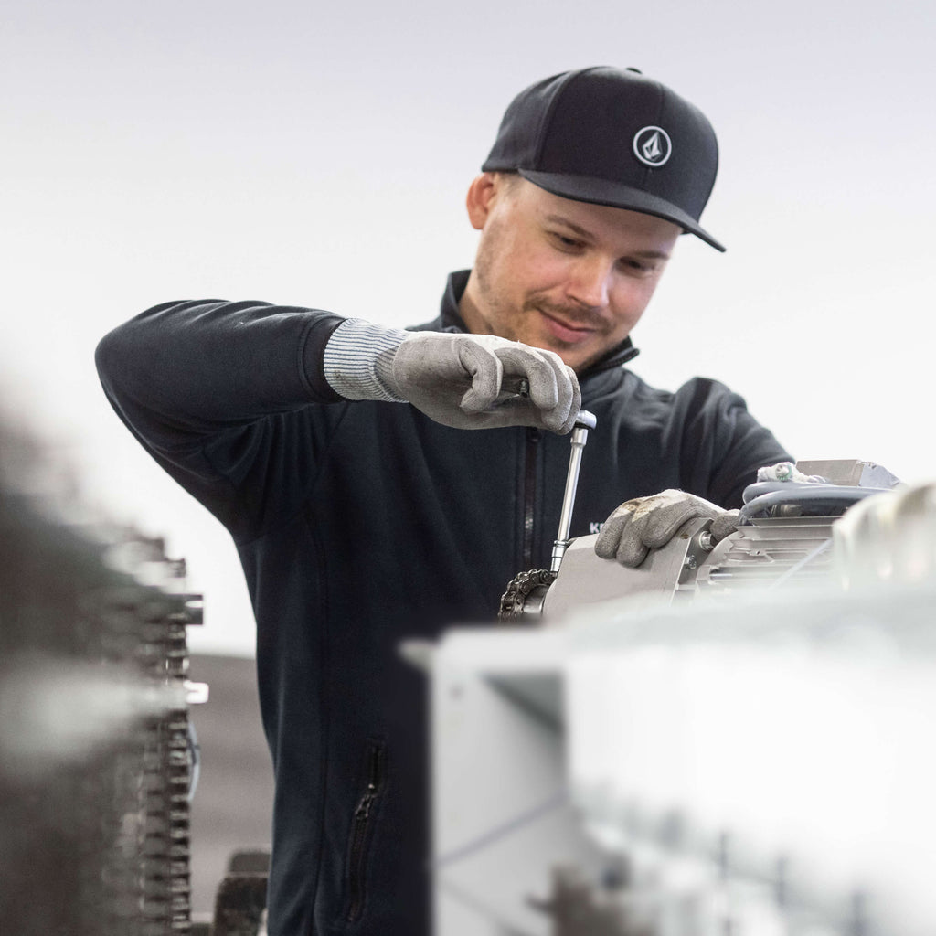 Ein Mann trägt eine schwarze Mütze und graue Handschuhe, während er mit einem Schraubenschlüssel an einer Maschine arbeitet. 
