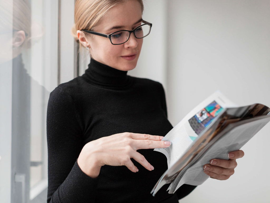Eine junge Frau mit schwarzem Rolli und Hornbrille steht lächelnd vor einem Fenster und blättert in einer Zeitschrift. Nur ihr Oberkörper, der sich unscharf im Fensterglas spiegelt , ist zu sehen.