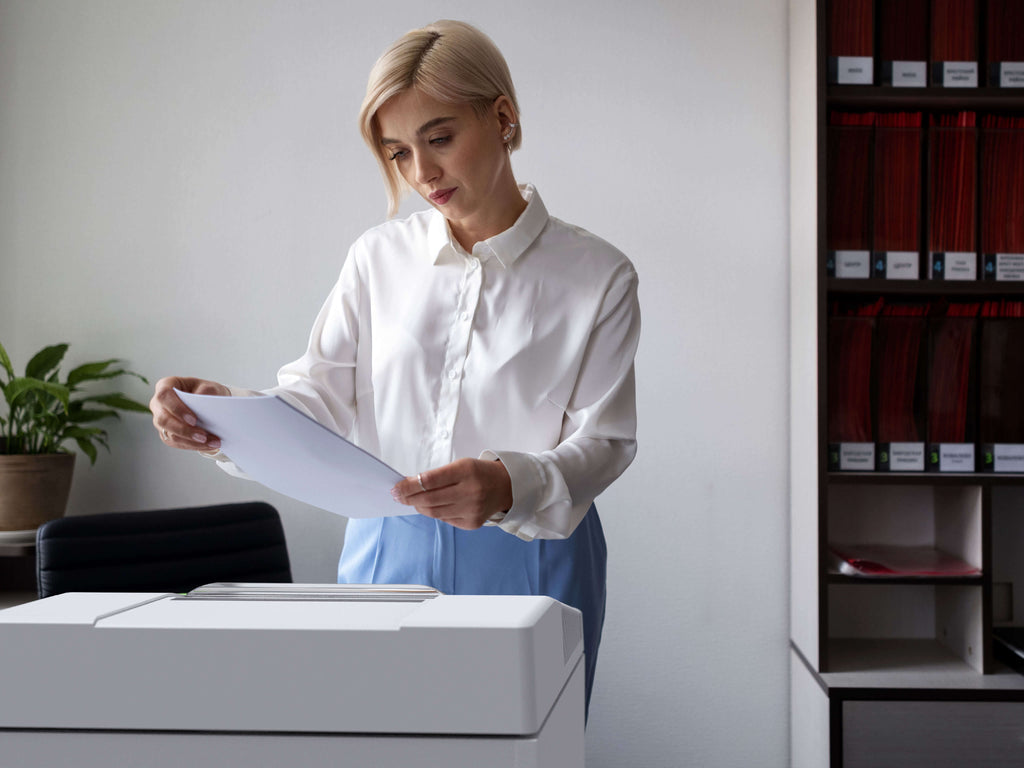 Eine Frau mit kurzen blonden Haaren, von vorne sichtbar, trägt eine weiße Hemdbluse und eine blaue Hose. Sie steht in einem Büro an einem IDEAL Aktenvernichter und betrachtet mit ernstem Blick ein Blatt Papier. Im Hintergrund befinden sich angeschnitten Regale mit Mappen und Büromaterial sowie eine Pflanze.