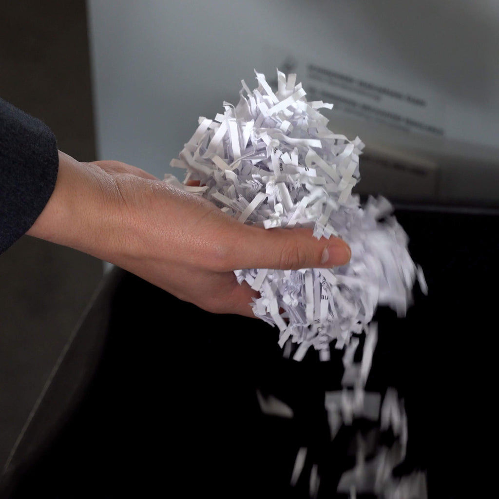 Eine Hand hält ein Häufchen geshreddertes weißes Papier, das gerade aus einem unscharf fotografierten IDEAL Schnittgutbehälter entnommen wurde. 