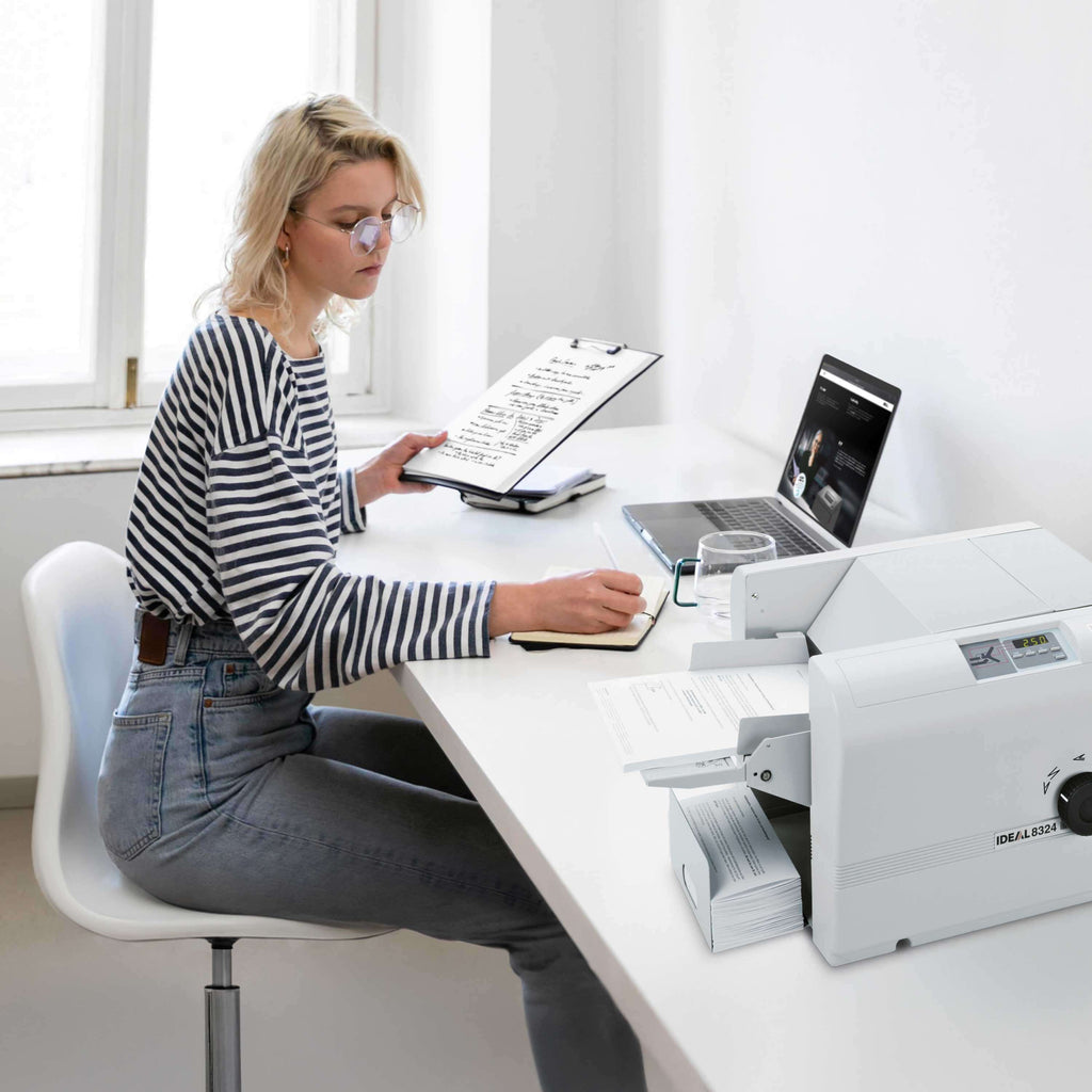 Eine junge Frau mit Brille, gestreiftem Langarm-Shirt und Jeans sitzt an einem Schreibtisch. Sie arbeitet mit einem Laptop und Dokumenten und macht sich Notizen mit einem Stift, neben ihr auf dem Tisch steht eine IDEAL 8324 Falzmaschine mit gefalzten und zu falzenden Briefen.