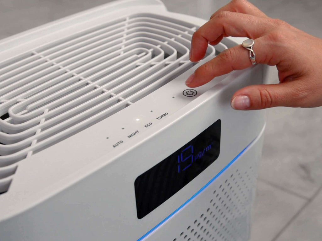 Eine Hand drückt auf die Ventilatorstufen-Bedientaste des Luftreinigers IDEAL AP25, der ausschnitthaft von oben fotografiert wurde. An der Oberseite und vorne sieht man die Luftauslässe, vorne oben ein digitales Display und diverse LED-Statusanzeigen (AUTO / NIGHT / ECO / TURBO). Ein horizontal verlaufendes Licht leuchtet in blau.