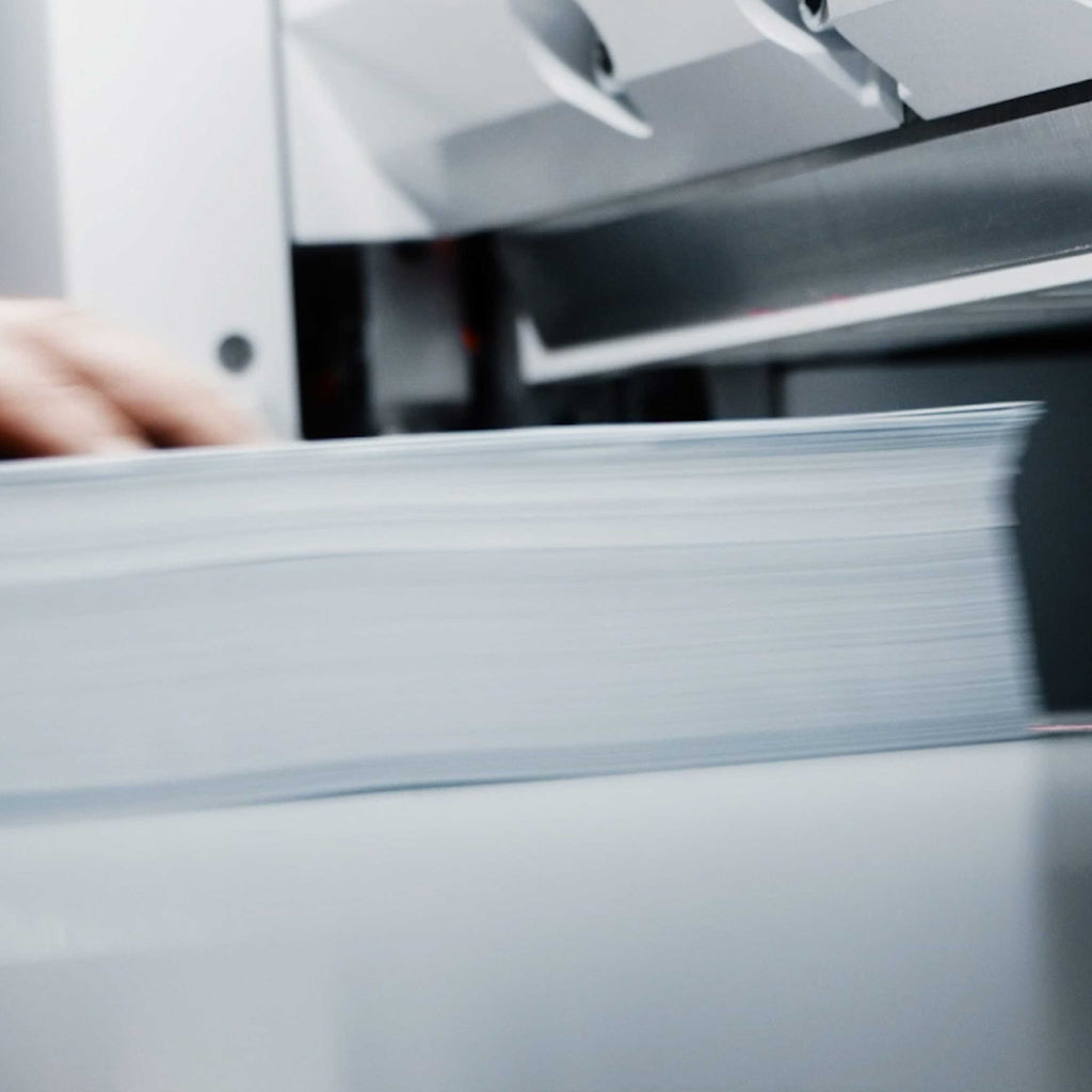 Unscharf fotografiertes Szenebild eines angeschnitten dargestellten Papierstapels, der in Nahaufnahme auf dem Tisch eines IDEAL Stapelschneiders liegt. Im Hintergrund oben das Schneidemesser.