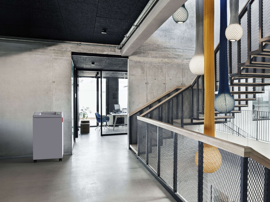 Modernes Treppenhaus mit Betonwänden und -böden, einer Treppe mit Metallgeländer und runden Hängelampen. Links steht ein IDEAL 2604 Büro-Aktenvernichter, durch Glastüren fällt der Blick auf ein Büro mit großen Glasfronten.