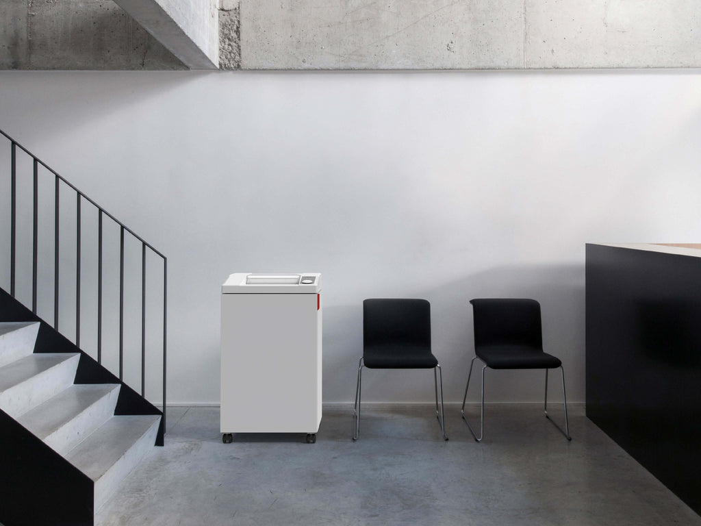 Minimalist reception area with concrete floor and two black chairs in front of a white wall, next to an IDEAL 3104 office shredder and a black reception desk. On the left, a staircase with metal railings leads upwards. 