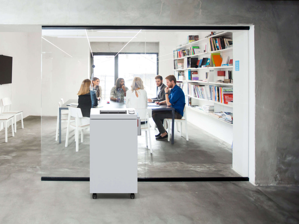 In einem modernen Konferenzraum mit weißen Möbeln, Glaswänden und Bücherregalen sitzt eine Gruppe um einen Tisch. Ein IDEAL 3105 Büro-Aktenvernichter mit automatischem Öler steht im Flur, direkt vor einem großen Glaspaneel im Vordergrund. 