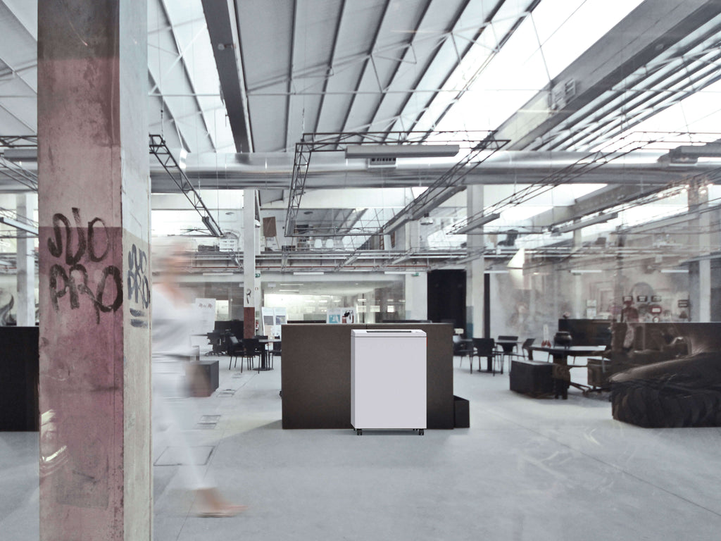 Modernes Großraumbüro im Industriedesign mit zentral platziertem IDEAL 4001 Büro-Aktenvernichter vor einem niedrigen grauen Trennelement. Transparente Deckenplatten lassen Tageslicht herein. Graffiti ziert einen Betonpfeiler, an dem eine Frau in Bewegungsunschärfe vorbeigeht.