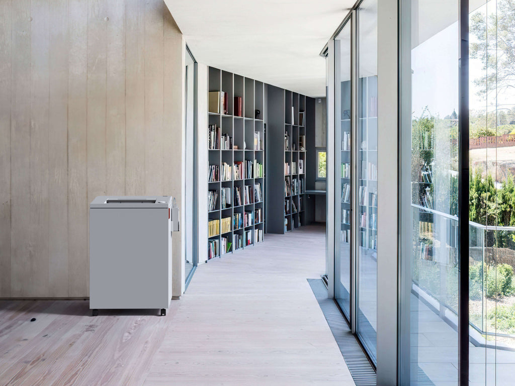 Modernes, lichtdurchflutetes, fast wie eine Bibliothek anmutendes Büro-Interieur mit Betonwand und Holzböden. Rechts eine große Glasfront, links mit Büchern gefüllten Regale entlang der geschwungenen Wand. Vorne links steht ein IDEAL 4003 Büro-Aktenvernichter.