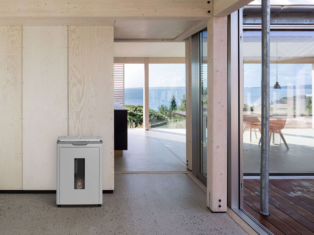 Modern interior with wooden walls, overlooking large windows with sea views. An IDEAL Shredcat autofeed shredder 8285 blends into the minimalist design on the left. Sunlit tables and chairs can be seen, with a terrace to the right.