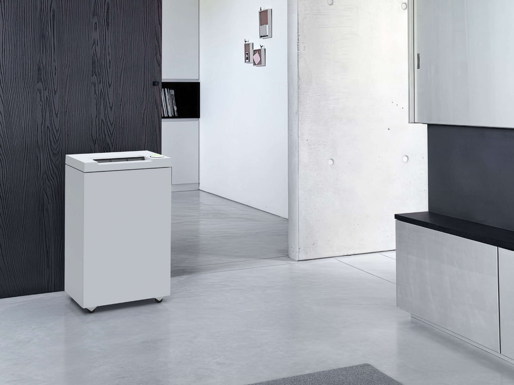 A modern, minimalist hallway room with light gray concrete floors and walls. On the left in the foreground is a light gray IDEAL 2465 desktop shredder in front of a dark wooden wall. The view leads into a room with small works of art on the wall and a clean cupboard.