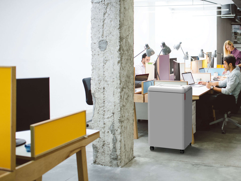 Modern open-plan office in industrial design with concrete pillars, concrete floor, low yellow and white partitions. People work on laptops at a long communal table. An IDEAL 2503 office shredder right next to the table ensures data protection.