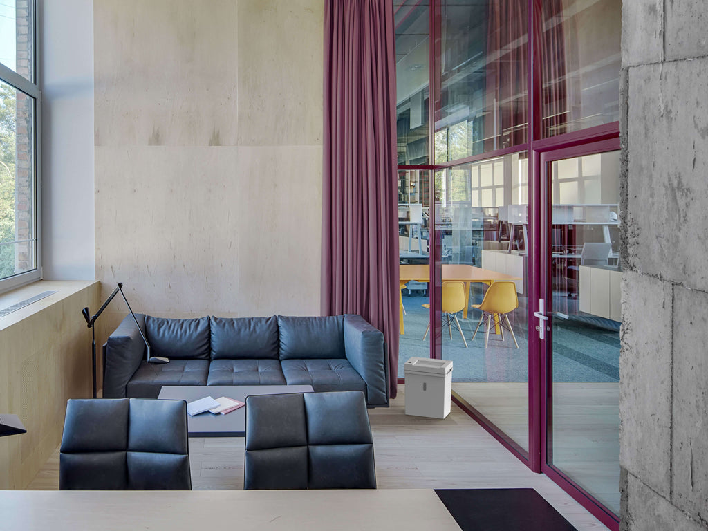 Modernes Einzelbüro mit Betonwänden, abgetrennt durch hohe Glasflächen mit raumhohem Vorhang und Tür von einem offenen Großraumbüro. Ein schwarzes Ledersofa steht direkt am Fenster, daneben steht ein IDEAL Shredcat 8200 Schreibtisch-Aktenvernichter. Helle Holz- und Betonelemente dominieren den Stil.