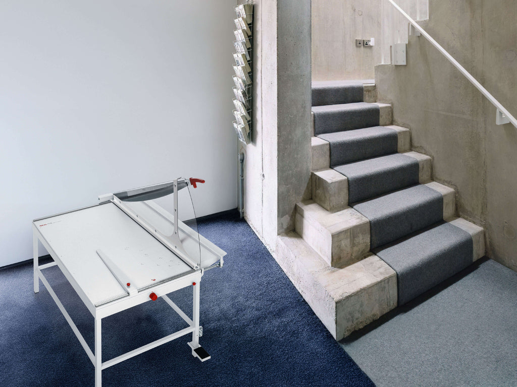 Moderner Treppenbereich mit einem großformatigen Profi-Hebelschneider IDEAL 1110 mit Untergestell  auf einem dunklen Teppich. Daneben führen mit Teppich belegte Stufen die Betontreppe hinauf. An der Betonwand hängt ein schmales, vertikales Display-Regal mit Broschüren.
