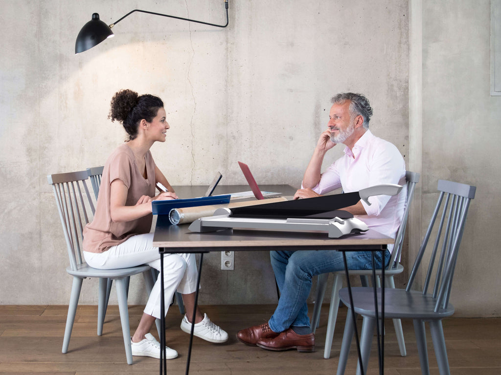 Eine Frau im T-Shirt und ein Mann in einem weißen Hemd sitzen mit Laptops an einem Tisch und lächeln sich an. Auf dem Tisch steht ein IDEAL 1142 Hebelschneider. Das Licht der Wandlampe fällt auf Holzböden und Betonwände.