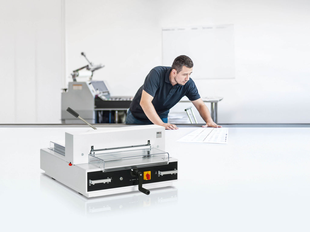 Ein Mann im Poloshirt prüft einen Druckbogen auf einem großen weißen Tisch. Im Vordergrund steht ein elektrischer Papier-Stapelschneider IDEAL 4315, ein kompaktes Tischgerät. Der Hintergrund ist verschwommen und zeigt eine Digitaldruckmaschine.