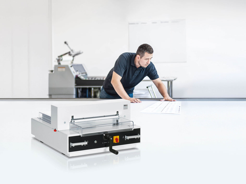 Ein Mann im Poloshirt prüft einen Druckbogen auf einem großen weißen Tisch. Im Vordergrund steht ein elektrischer Papier-Stapelschneider IDEAL 4350, ein kompaktes Tischgerät mit automatischer Pressung. Der Hintergrund ist verschwommen und zeigt eine Digitaldruckmaschine.