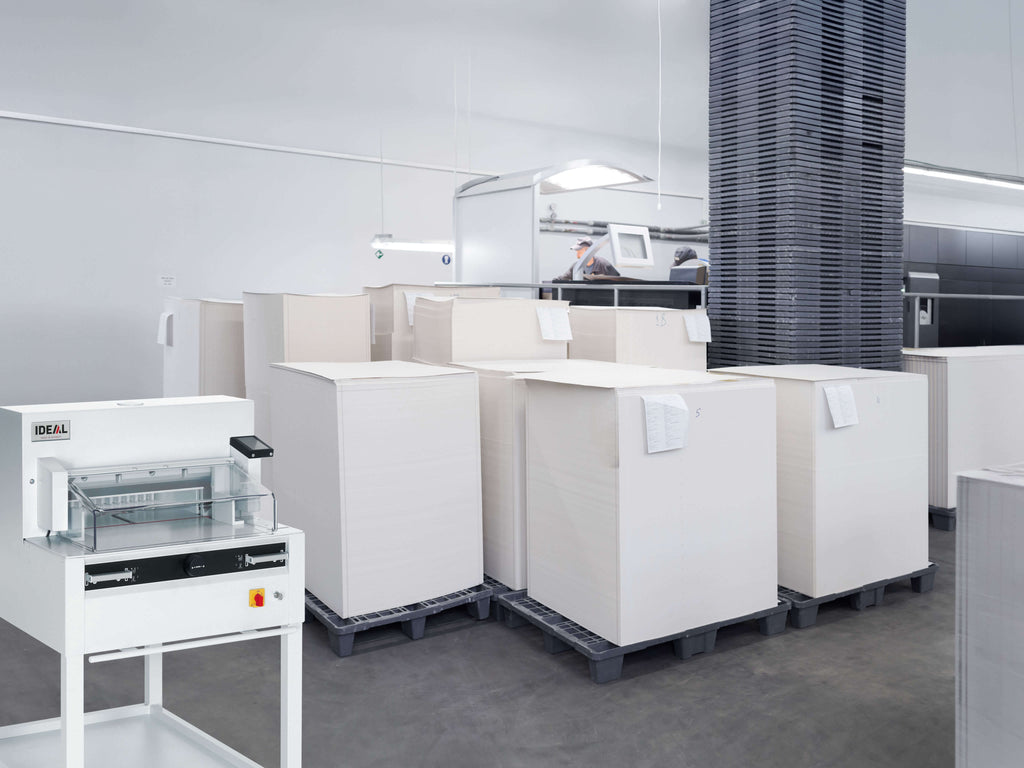 Ein geräumige, moderne Druckerei mit mehreren Paletten voll weißer Papierbögen. Im Vordergrund ein IDEAL 4855 Stapelschneider zum Beschneiden von Druckbögen. Im Hintergrund des gut beleuchteten Raums mit Betonboden steht eine moderne Druckstraße.
