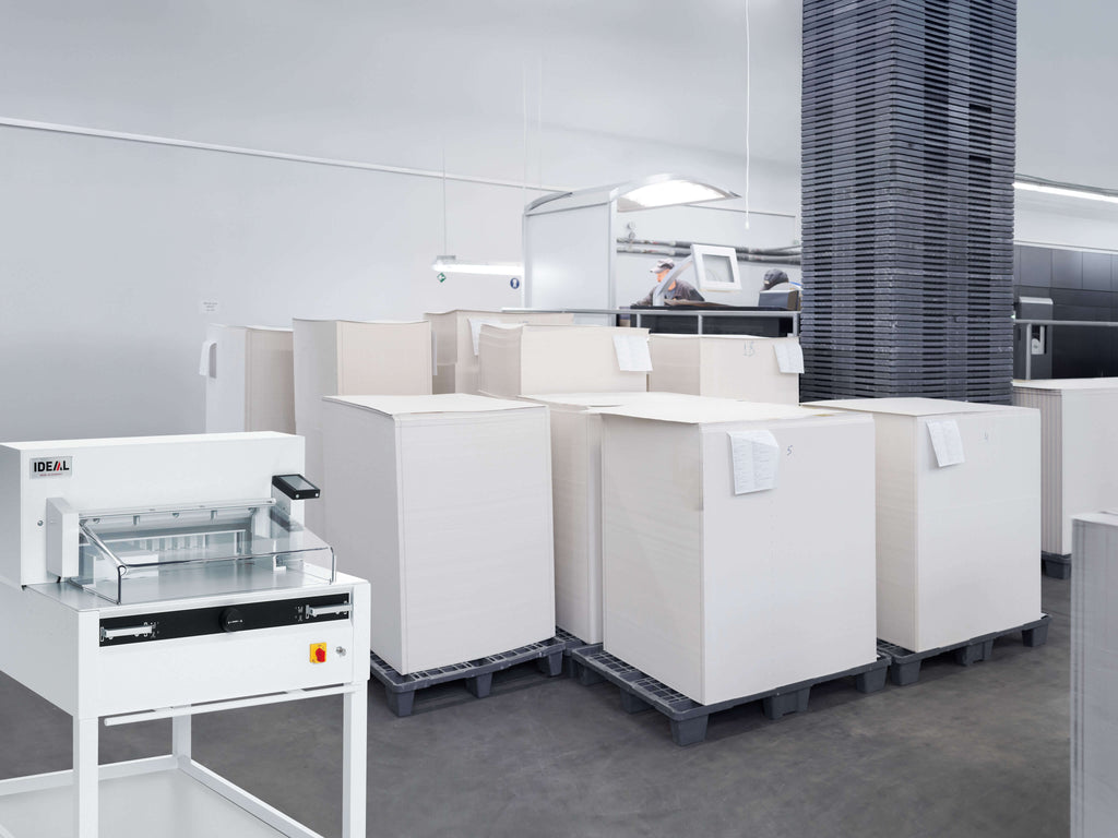 Ein geräumige, moderne Druckerei mit mehreren Paletten voll weißer Papierbögen. Im Vordergrund ein IDEAL 5255 Stapelschneider zum Beschneiden von Druckbögen. Im Hintergrund des gut beleuchteten Raums mit Betonboden steht eine moderne Druckstraße.