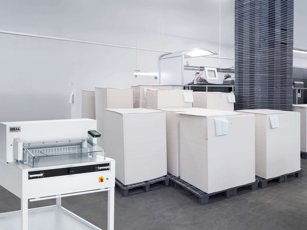 Ein geräumige, moderne Druckerei mit mehreren Paletten voll weißer Papierbögen. Im Vordergrund ein IDEAL 6655 Stapelschneider zum Beschneiden von Druckbögen. Im Hintergrund des gut beleuchteten Raums mit Betonboden steht eine moderne Druckstraße.