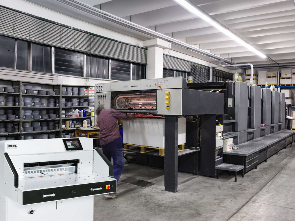 In einer Druckerei arbeitet eine Person an einer großen Druckstraße mit Farb-Druckwerken. Regale mit Farbdosen säumen die Wände, Leuchtstoffröhren erhellen den Raum im Industriestil. Im Vordergrund ein IDEAL 7260 Papier-Stapelschneider mit Lichtschranke und Lufttisch zum Beschneiden von Druckbögen.