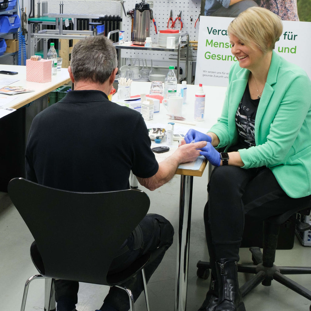 Frau in grüner Jacke und blauen Handschuhen misst den Blutzucker eines Mannes in schwarzem T-Shirt an einem Tisch mit medizinischen Utensilien.