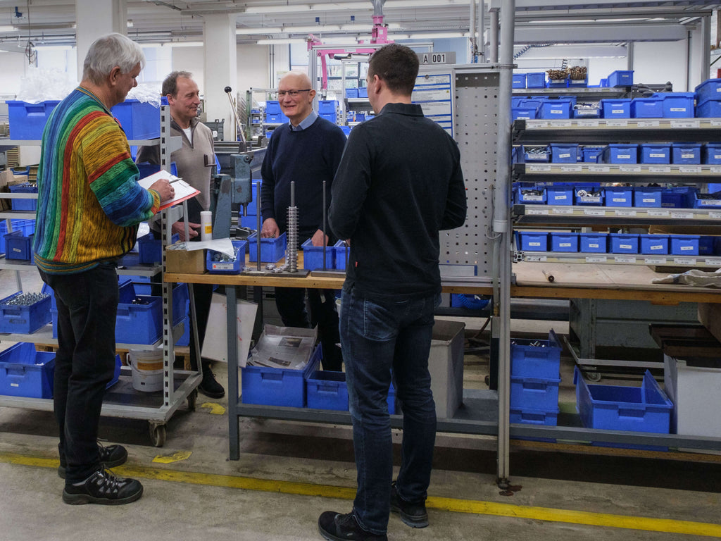 Vier Männer stehen und unterhalten sich um eine Werkbank in einer Fabrik oder Werkstatt, umgeben von Regalen mit blauen Behältern und Werkzeugen. Ein Mann macht sich Notizen auf einem Klemmbrett. 