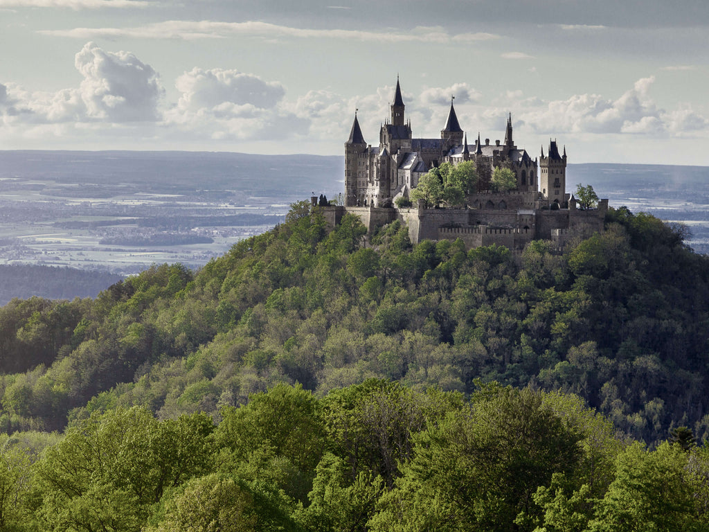 Burg Hohenzollern auf bewaldetem Berg mit mehreren Türmen und Zinnen, im Hintergrund weite Landschaft unter bewölktem Himmel.