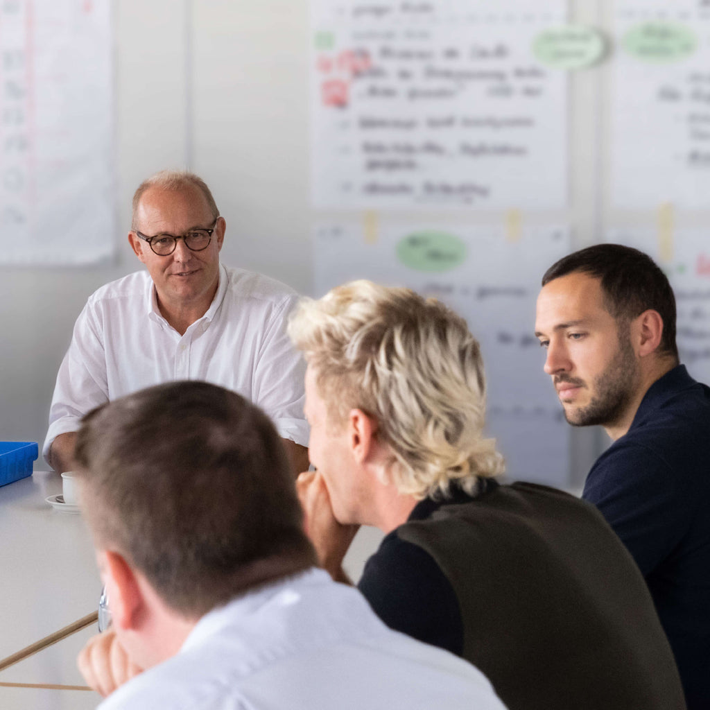 Vier Männer sitzen in einem hellen Raum an einem Tisch und diskutieren, im Hintergrund hängen Tabellen und Notizen an der Wand. Ein Mann mit Brille scheint das Gespräch zu leiten. 