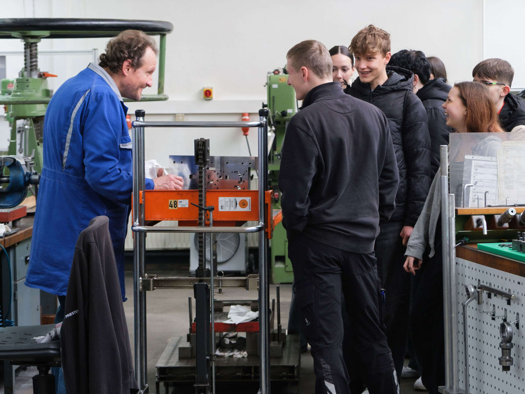 Ein Mann in einem blauen Kittel erklärt einer Gruppe aufmerksamer junger Leute in einer mit Geräten gefüllten Werkstatt Maschinen. 