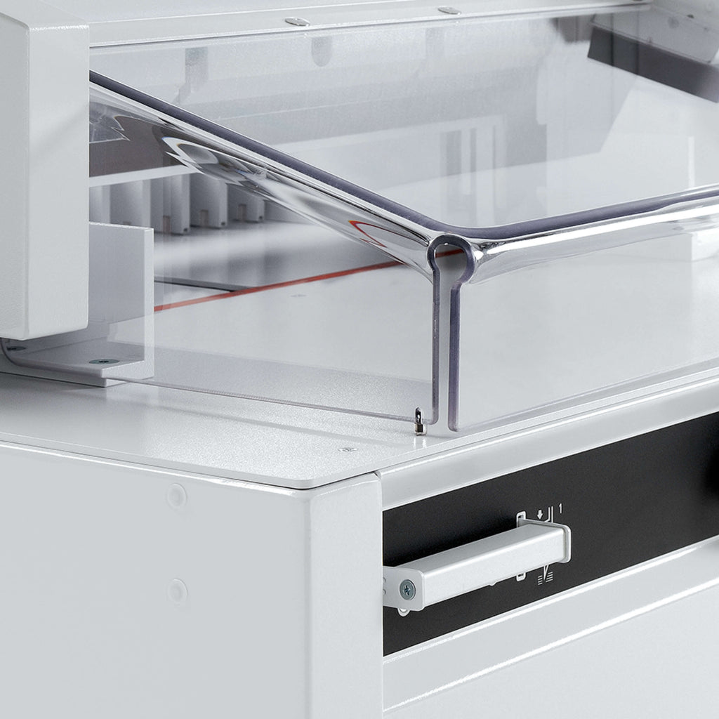 Detailansicht eines elektrischen IDEAL Papier-Stapelschneiders. Zu sehen die transparente Sicherheits-Haube. Der Blick fällt auf einen Teil des metallenen Maschinentischs, darunter ist ein Schnittauslöse-Taster zu sehen.