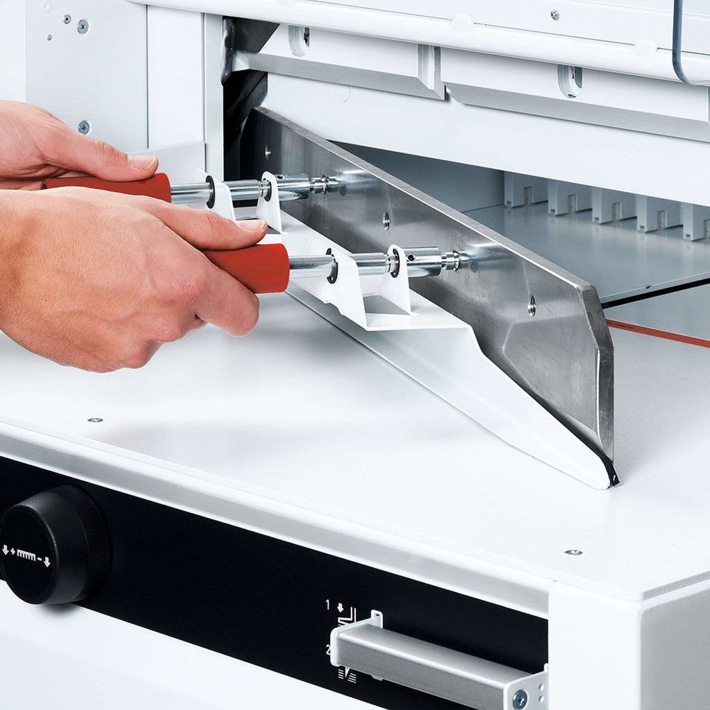 Nahaufnahme einer für den Bediener sicheren Messerwechsel-Szene bei einem IDEAL Stapelschneider 4855, 5255 oder 6655. Zwei Hände halten ein Werkzeug mit dem Stapelschneider-Messer. Die Hände sind geschützt. Man sieht noch einen Teil der Maschine mit elektronischem Handrad und EASY Cut-Schnittauslöser.