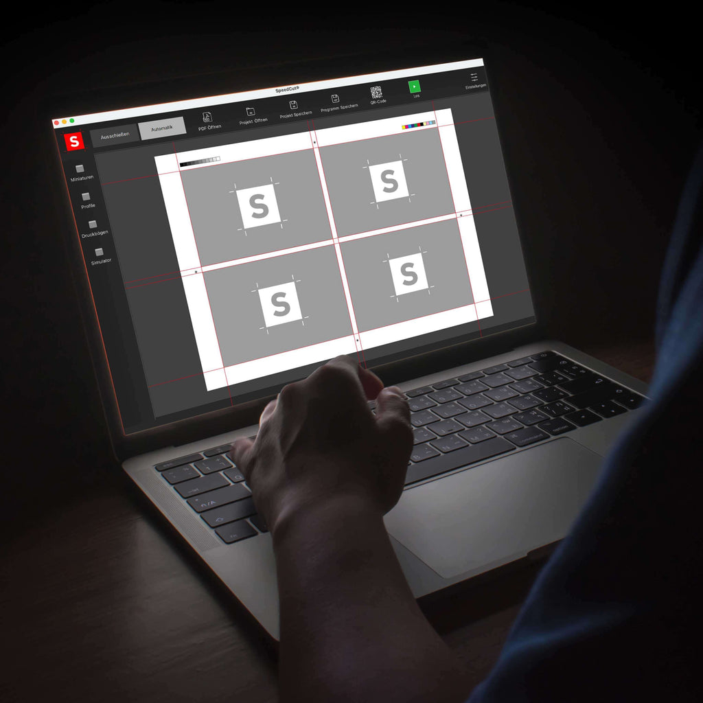 Von hinten fotografierter Bediener, unscharf im Dunkeln, dessen linke Hand auf einer Laptop-Tastatur liegt. Auf dem Monitor ist die Bedienoberfläche des Schnitt-Automatisierungs-Programms Speedcut für die IDEAL Stapelschneider THE56 oder THE68 zu sehen, mit einem Schnittbogen, der 4 Nutzen zeigt. Das Programm bietet eine automatische Schnittmarkenerkennung.
