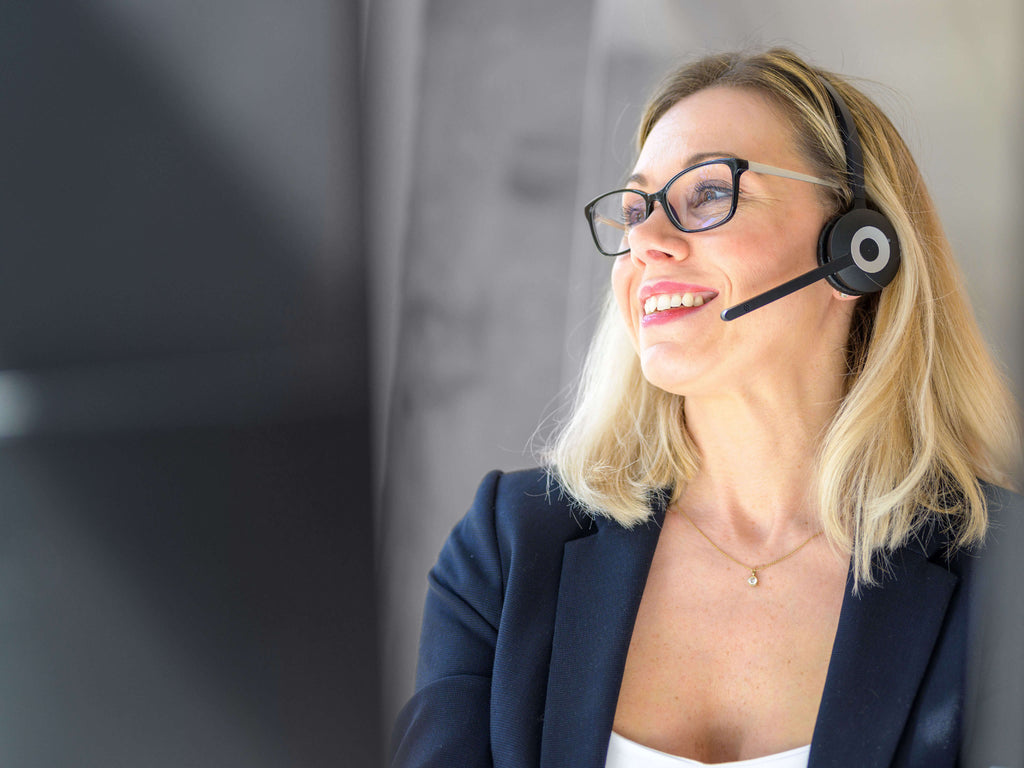 Eine junge Frau, schickt gekleidet in dunkelblauem Blazer und weißem Top, mit Brille und Headset, von vorne fotografiert, hört lächelnd zu. Der Hintergrund st unscharf-grau. Das Bild symbolisiert den Telefon-Kundendienst von IDEAL.