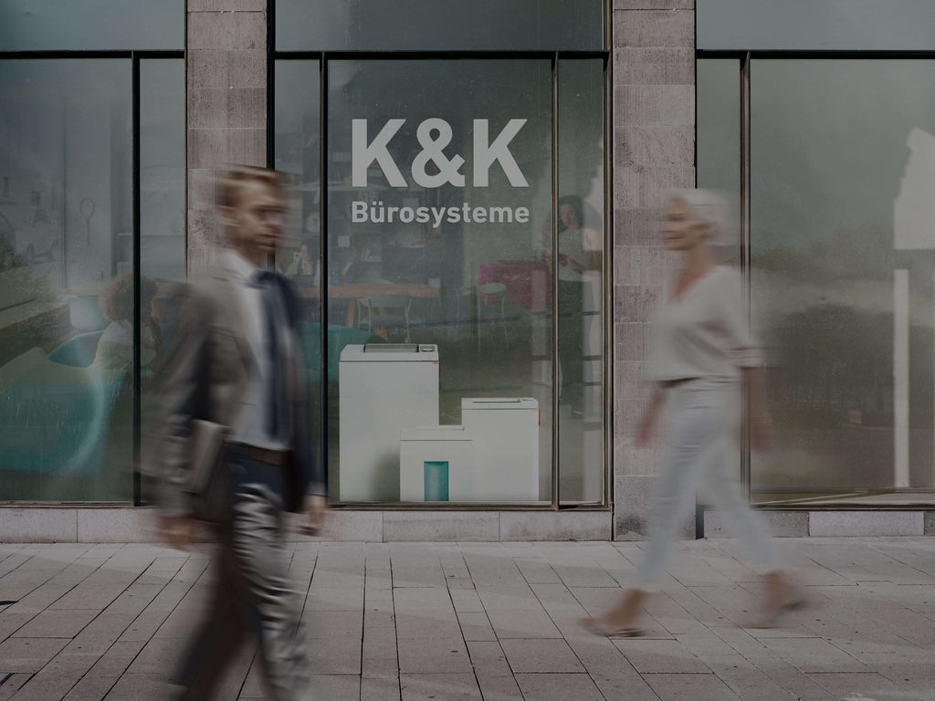 Schaufenster mit der Aufschrift 'K&K Bürosysteme'. Im Fenster sind Büromöbel und IDEAL Aktenvernichter sichtbar. Zwei Personen gehen vorbei.