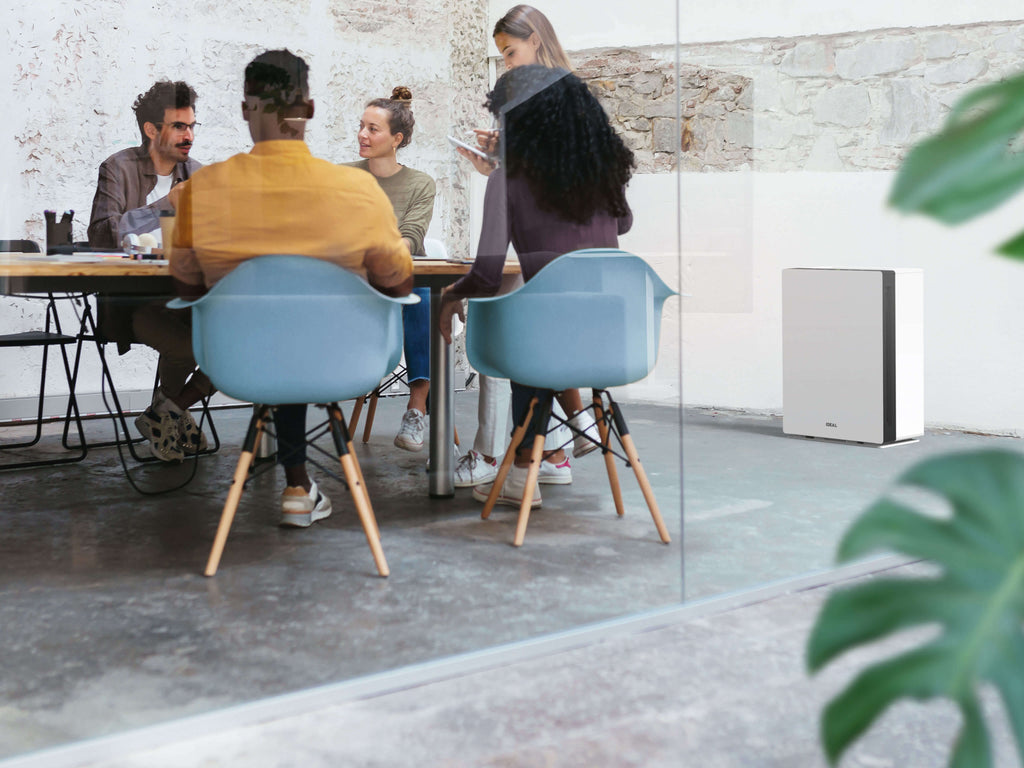 Fünf Personen sitzen oder stehen in einem modernen Besprechungsraum mit Glaswänden und Betonboden um einen Holztisch und diskutieren. Der Raum ist minimalistisch eingerichtet, mit hellblauen Stühlen und einer Pflanze. Ein IDEAL AP60 PRO Luftreiniger sorgt für saubere Luft.