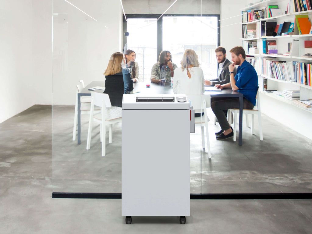 In einem modernen Konferenzraum mit weißen Möbeln, Glaswänden und Bücherregalen sitzt eine Gruppe um einen Tisch. Ein IDEAL 3105 Büro-Aktenvernichter mit automatischem Öler steht im Flur, direkt vor einem großen Glaspaneel im Vordergrund. 