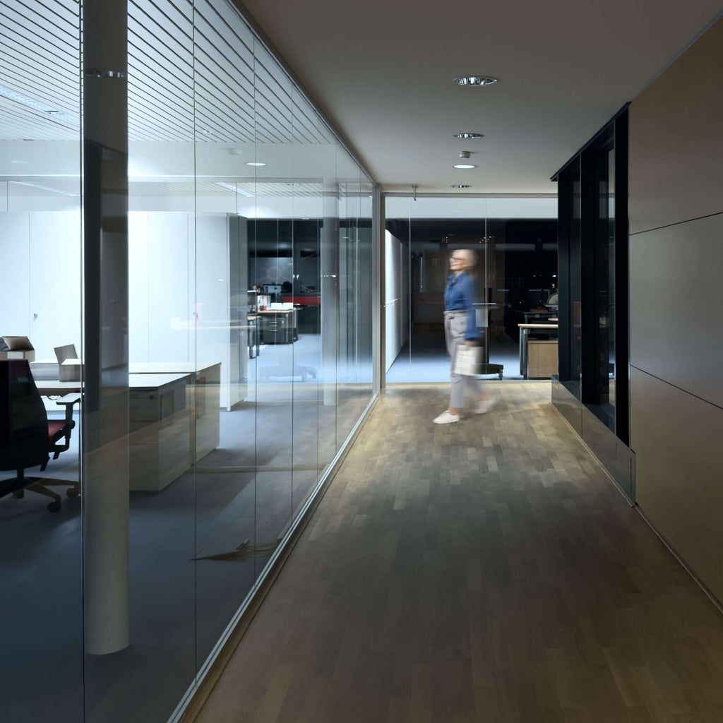 Eine Person in blauer Kleidung geht durch einen modernen Büroflur mit Glaswänden innerhalb des Firmengebäudes von IDEAL. Im Hintergrund sind Büromöbel sichtbar. Die Szene ist schwach beleuchtet.