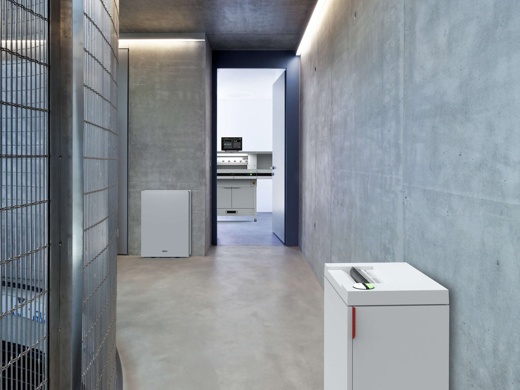 Ein moderner Raum mit Betonwänden. Links steht ein IDEAL Luftreiniger, rechts ein IDEAL Aktenvernichter. Im Hintergrund ist eine offene Tür zu einem Raum mit einem IDEAL Stapelschneider.